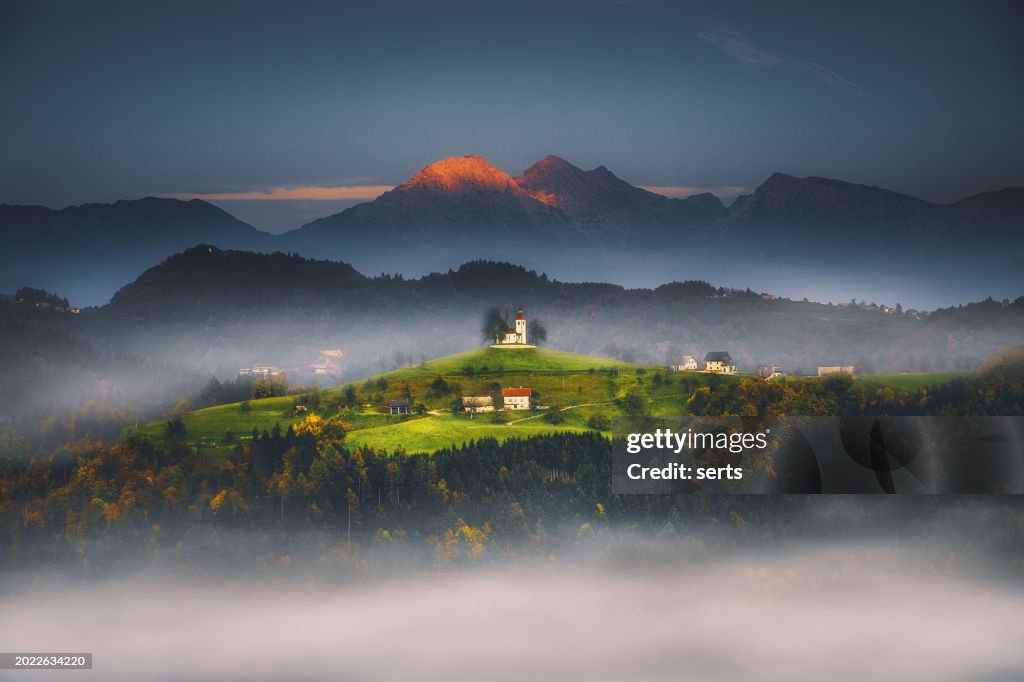 Vista del paisaje con la iglesia de Sveti Tomaz (Santo Tomás) o la iglesia (Cerkev Sveti Tomaz) en un hermoso amanecer en otoño, cerca de Skofja Loka en la región de Alta Carniola, Eslovenia, Europa