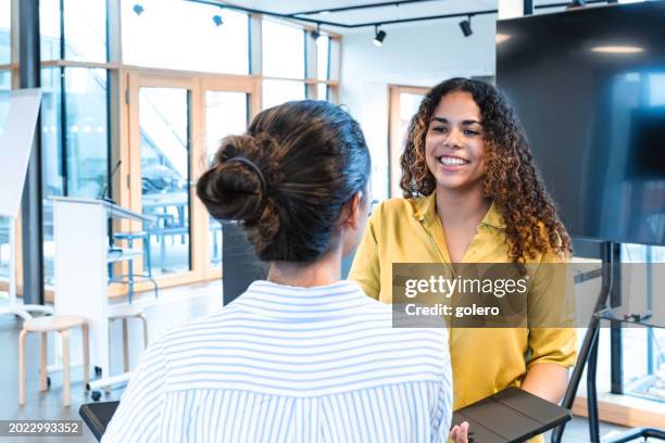 junge lächelnde frau mit digitalem tablet, die mit einer anderen frau spricht - woman 20 summit in berlin stock-fotos und bilder