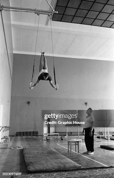 Gymnast performs on the still rings suspended from a wall-mounted bracket, watched by a man below, in a gym in Moscow, Russia, Soviet Union, circa...