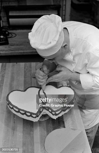 High-angle view as a patissier uses a piping bag to apply icing to a wedding cake in the shape of two love hearts, United States, 1963.