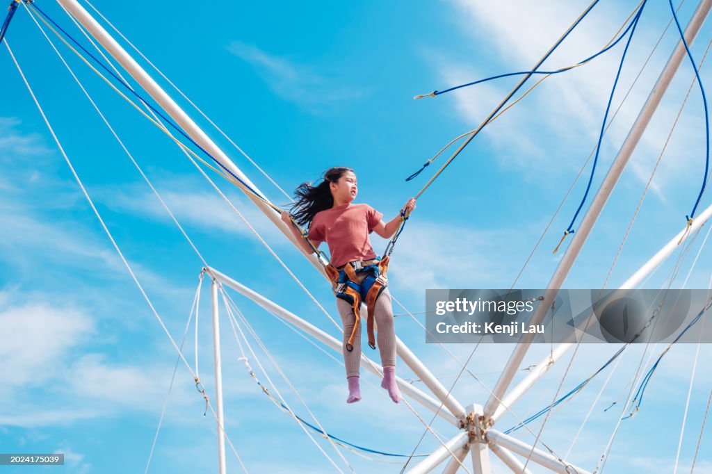 Happy little Asian girl in air secured in a rope jumping on trampoline on summer day against background of clear blue sky.