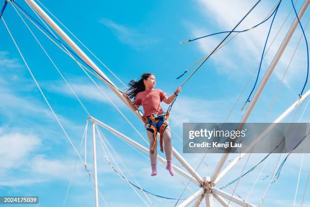 happy little asian girl in air secured in a rope jumping on trampoline on summer day against background of clear blue sky. - extreme sports kids stock pictures, royalty-free photos & images