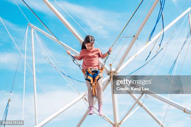 happy little asian girl in air secured in a rope jumping on trampoline on summer day against background of clear blue sky. - extreme sports kids stock pictures, royalty-free photos & images