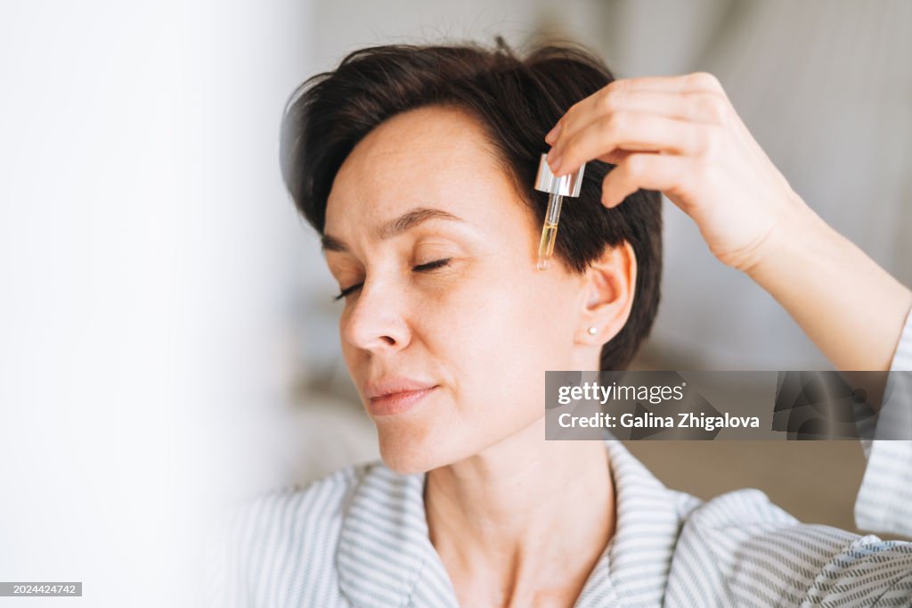 Young adult brunette woman with short hair applies oil to her face at home, morning skincare routine