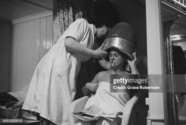 Staff helps a client sitting at a hair hood in the changing room at the Savoy, a women's Turkish bath on Duke of York Street, London, UK, 1951....