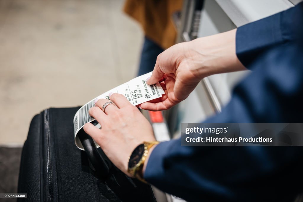 Close Up Of Airport Staff Member Attaching Tag To Passenger's Luggage