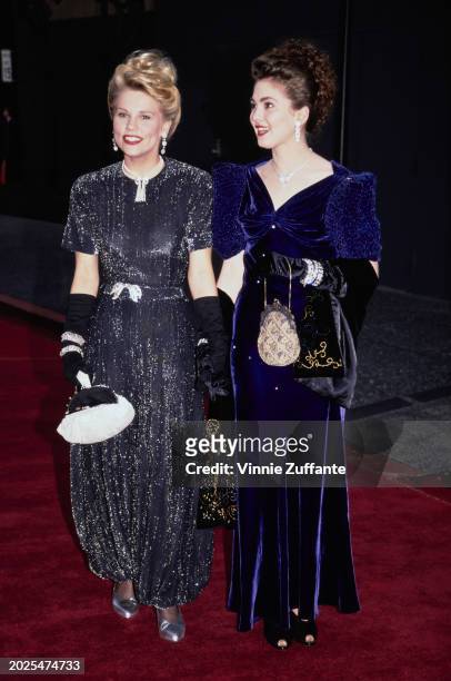 British actress Sammi Davis, wearing a short-sleeved grey evening gown with black evening gloves, holds hands with a guest, who wears a midnight blue...