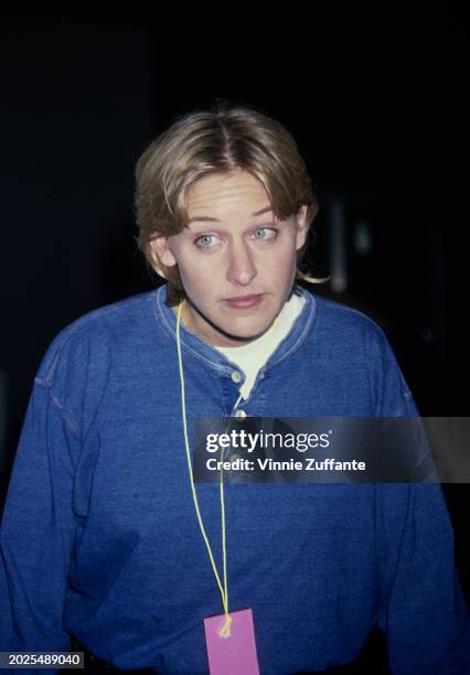 American television host and comedian Ellen DeGeneres, wearing a blue grandad shirt over a white crew-neck top, a pink card hanging from a lanyard...