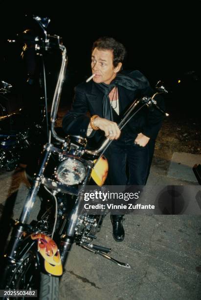 British actor and singer Michael Des Barres, wearing a black suit with a black scarf, poses beside a motorcycle at the IFP 'Rebel With A Cause'...