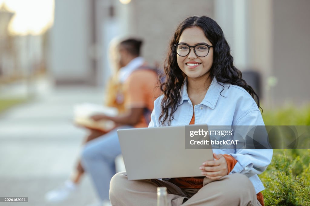 Portrait of a young Latin female student using laptop on the bench on campus