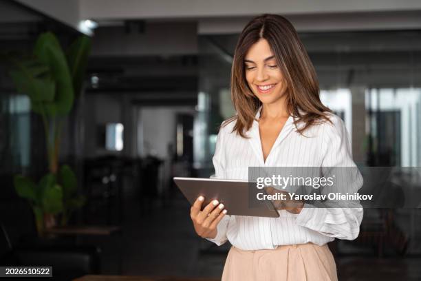 Latin Hispanic mature adult professional business woman looking at online trade app. European businesswoman CEO holding digital tablet using fintech tab application standing at workplace in office.