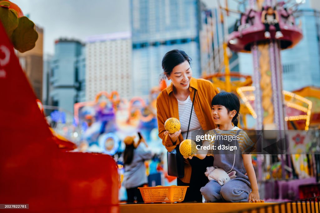 Joyful young Asian mother and lovely daughter playing games at the funfair to win a prize. Happy family having fun and enjoying spending time together at the carnival. Family lifestyle. Family fun time. Creating memorable moments