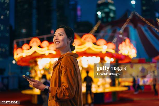 smiling young asian woman using smartphone in front of illuminated carnival booth at the funfair. having fun time at the amusement park at night. lifestyle and technology. games and entertainment - fairground stall stock pictures, royalty-free photos & images