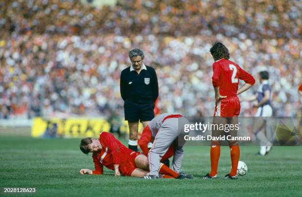 Referee Alan Robinson and Liverpool central defender Mark Lawrenson look on as Liverpool player-manager Kenny Dalglish is treated on the pitch for an...