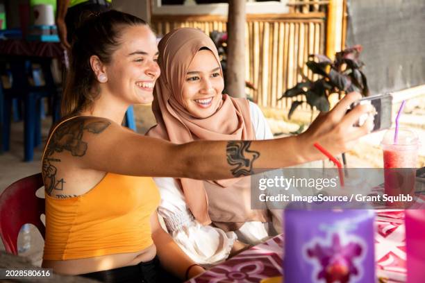 two young female friends one wearing a hijab sit together in a restaurant playing with their phones and taking selfies - tooth bonding stock pictures, royalty-free photos & images