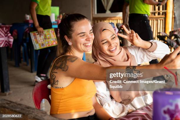 two young female friends one wearing a hijab, sit together in a restaurant playing with their phones and taking selfies - tooth bonding stock pictures, royalty-free photos & images