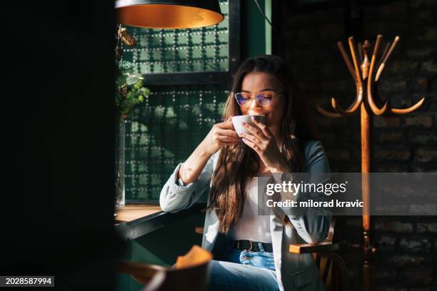 a young beautiful girl is sitting in a cafe by the window and enjoying her coffee. - view-through-restaurant-window stock pictures, royalty-free photos & images
