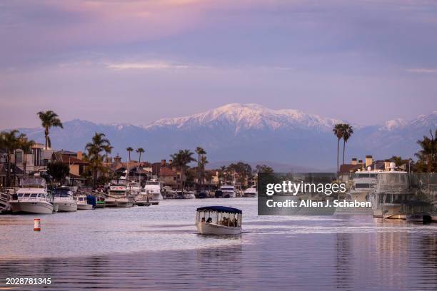 Huntington Beach, CA People in an electric boat take a scenic cruise as the setting sun illuminates clouds and the top of recently snow-capped San...