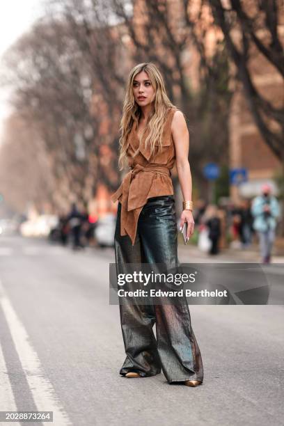 Guest wears a sleeveless brown low-neck suede double breasted jacket with integrated belt, blue and purple shiny painted flared denim pants / jeans ,...