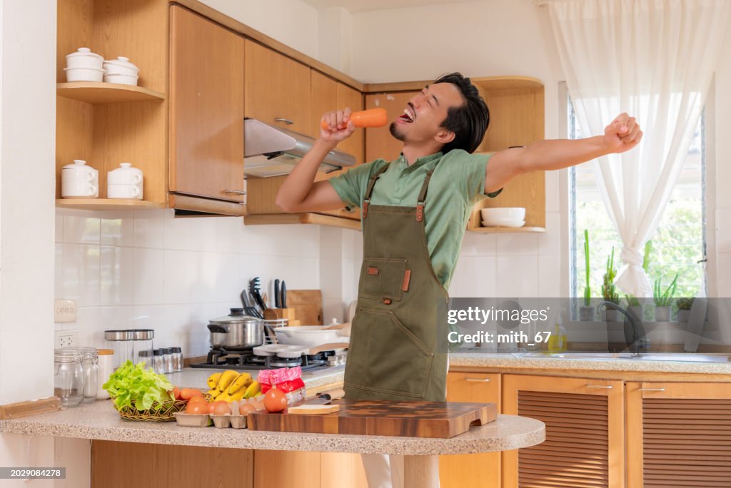 A funny Japanese mid-adult man dancing and singing in a kitchen while preparing his meals, using carrot as microphone, relaxing at home on weekends