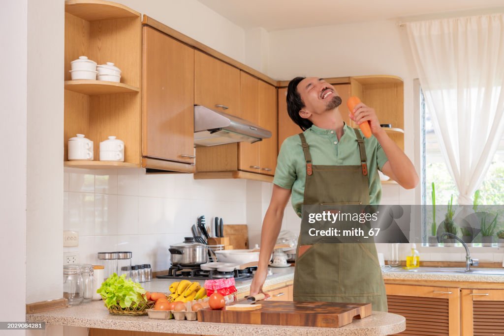 A funny Japanese mid-adult man dancing and singing in a kitchen while preparing his meals, using carrot as microphone, relaxing at home on weekends