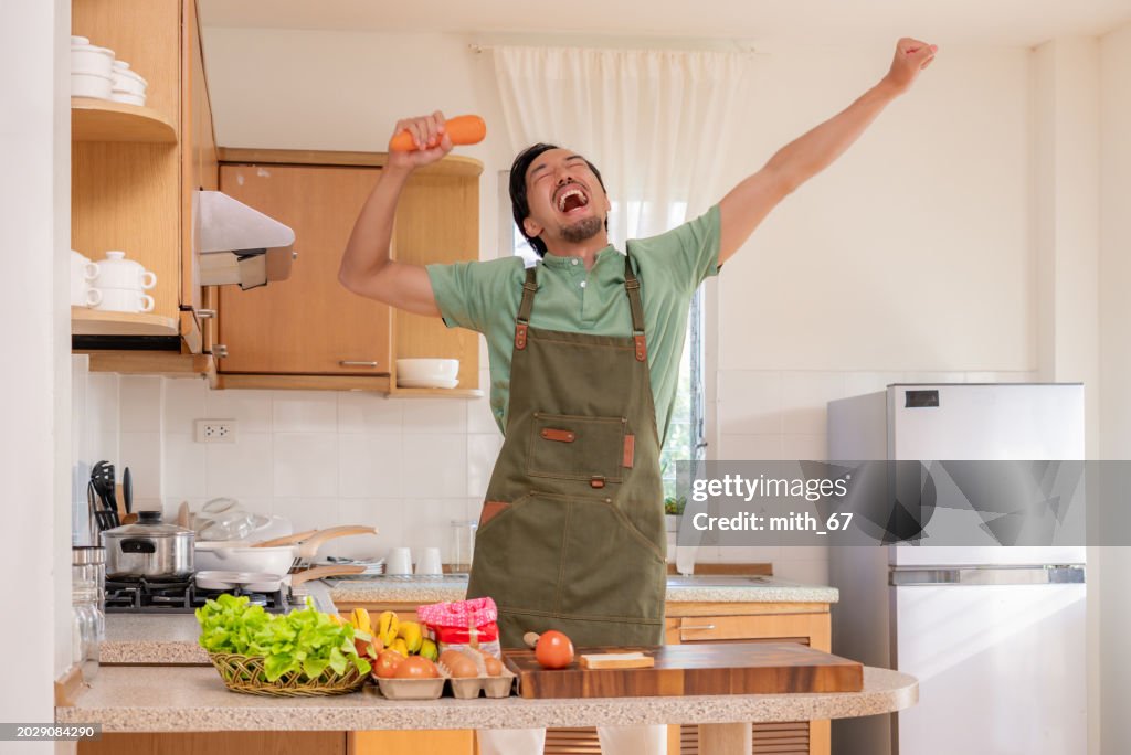 A funny Japanese mid-adult man dancing and singing in a kitchen while preparing his meals, using carrot as microphone, relaxing at home on weekends