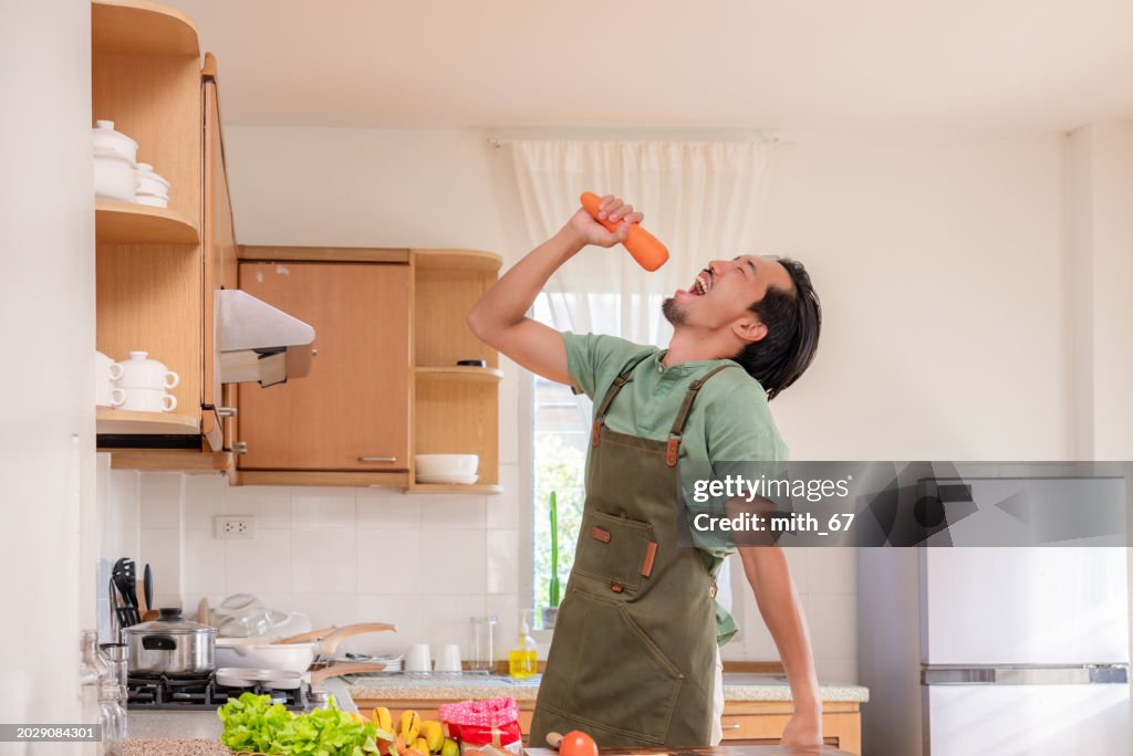 A funny Japanese mid-adult man dancing and singing in a kitchen while preparing his meals, using carrot as microphone, relaxing at home on weekends