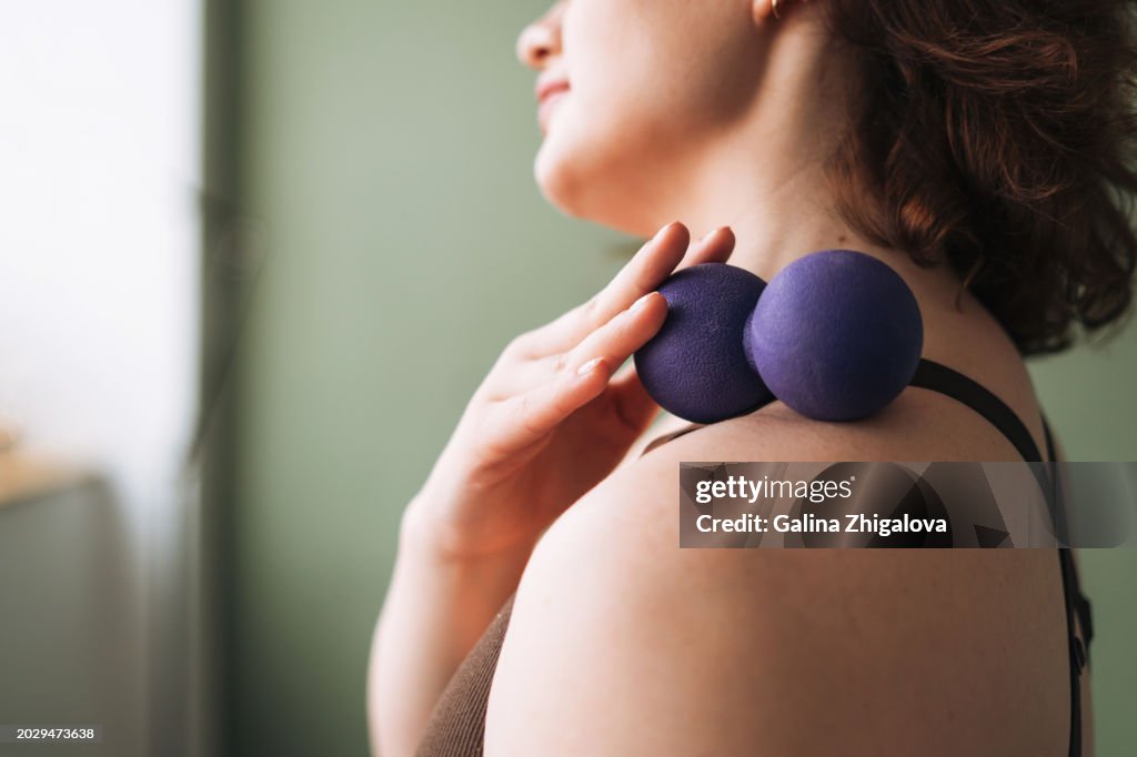 Brunette smiling young woman in sports uniform doing self-massage with massage ball after fitness exercises at home