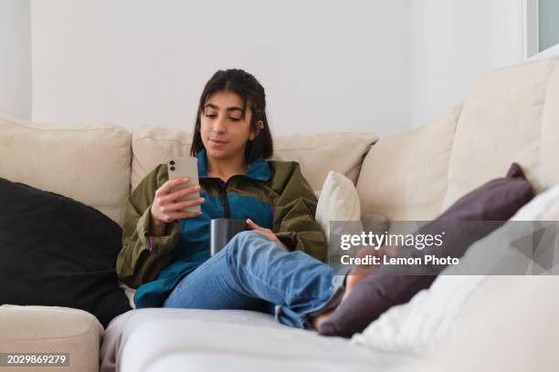 medium shot of a carefree and authentic generation z latino female taking a break sitting on a confortable sofa using a smartphone taking some hot drink at home on a sunny day - confortable imagens e fotografias de stock