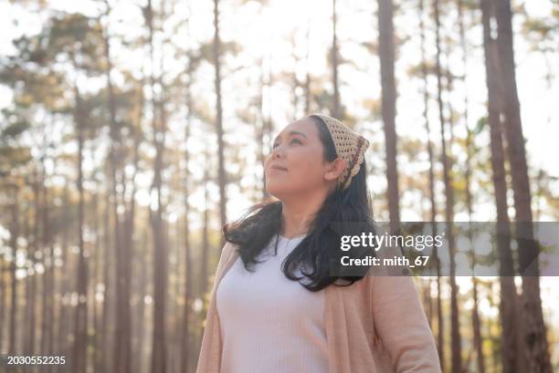 the asian mid-adult woman in white t-shirt and beige cardigan, surrounded by pine trees in the forest, is inhaling and feeling nature, exuding a positive and refreshing mood. - exuding stock pictures, royalty-free photos & images