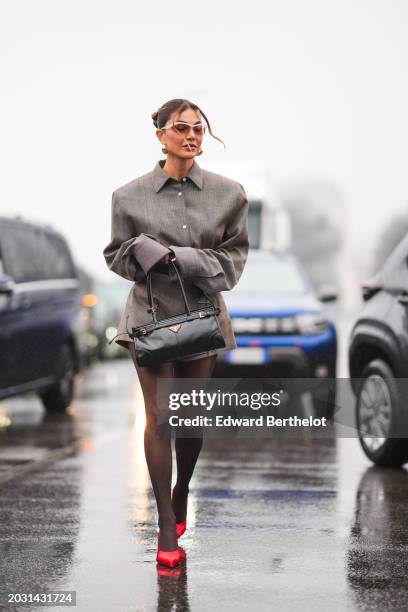 Negin Mirsalehi wears sunglasses, earrings, a gray oversized shirt with shoulder pads and long sleeves, a belt, flared pleated shorts, tights, a...