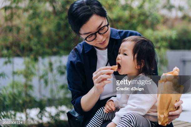 young asian mother sharing freshly made traditional street snack egg waffle to her lovely little daughter who sits on her lap at local food market in hong kong - hong kong community 個照片及圖片檔