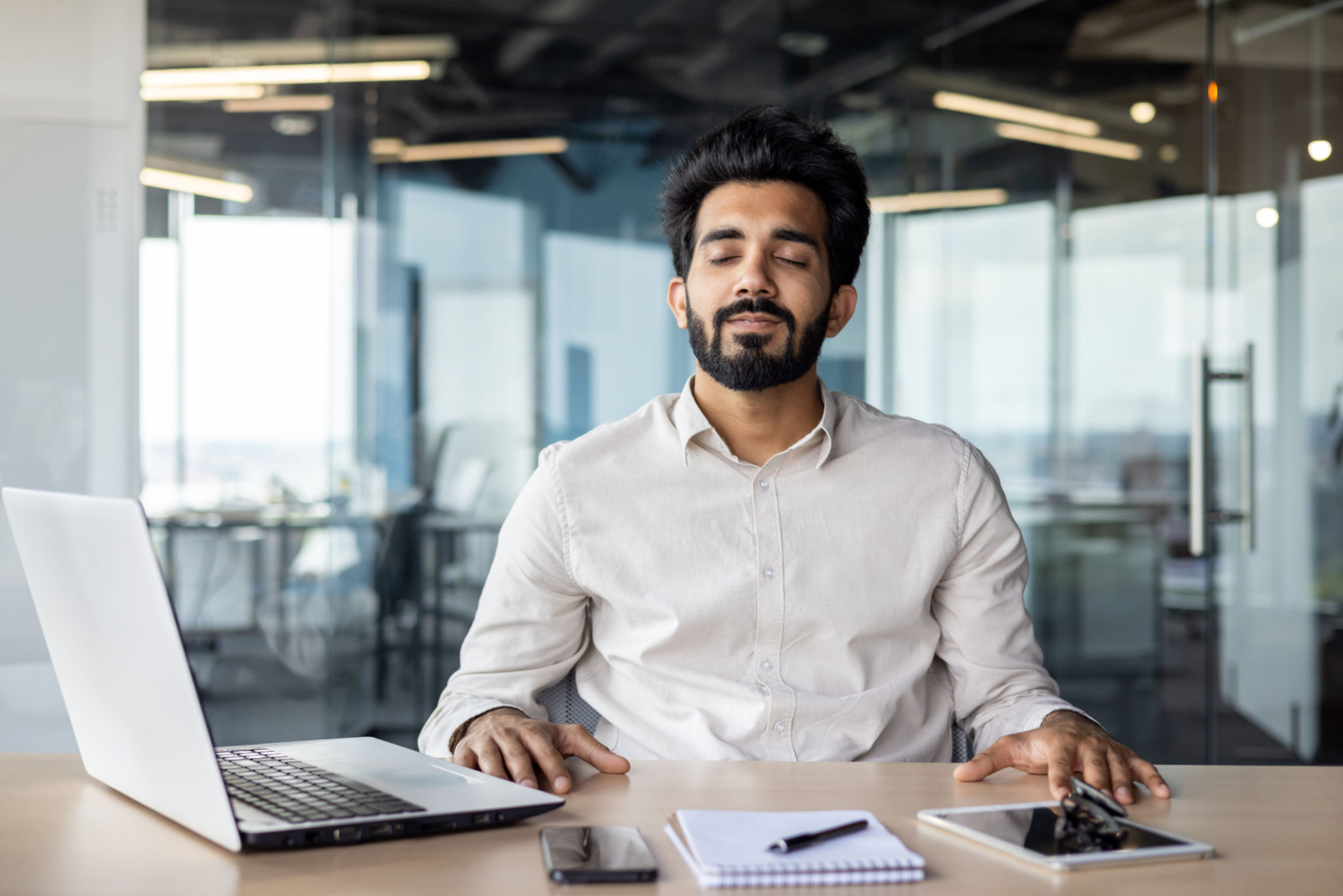 Relaxed young Indian man sitting in office at desk with laptop and documents and resting with closed eyes Relaxed young Indian man sitting in office at desk with laptop and documents and resting with closed eyes
