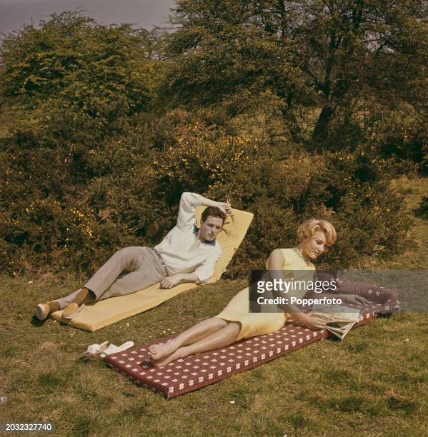 Young man and woman posed lying on sun loungers outdoors in a garden, she wears a yellow sleeveless dress and reads a magazine, England, 23rd June...
