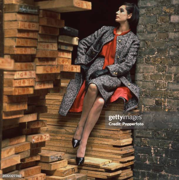 Female fashion model posed wearing a Chanel black and white herringbone coat with red lining, a matching skirt and red bow blouse, she sits on planks...