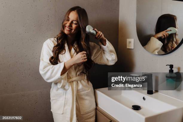 woman combing her beautiful hair with brush while standing near mirror in bathroom - thick women stock pictures, royalty-free photos & images