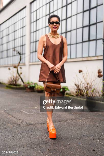 Tamu Mcpherson wears sunglasses, a golden chain necklace, a brown lustrous shiny silk low-neck sleeveless mini dress , a brown bag, orange platform...