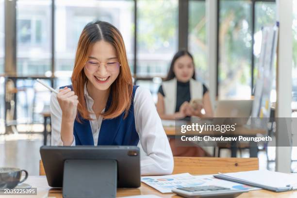young gen z woman working on her project using notebook computer at a co-working space smiling and clenching her fist in excitement while another woman working in the background - z com stock pictures, royalty-free photos & images