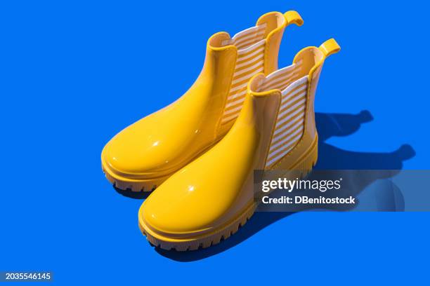 yellow rain boots with white and yellow striped elastic tape on blue background. concept of rain, footwear, boots, design, contrast, art and color. - zapatos-amarillos fotografías e imágenes de stock