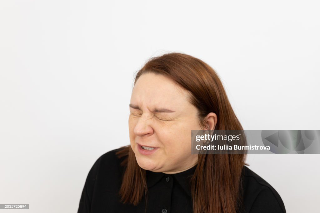 Expressive portrait of redhead woman, standing in front of white background with eyes closed and face contorted in grimace