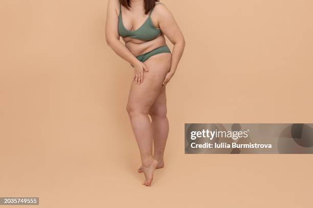 unrecognizable chubby woman in bikini slightly bending over, delicately touching skin on thigh, massaging limb, exuding self-love in studio setting. - exuding stock pictures, royalty-free photos & images