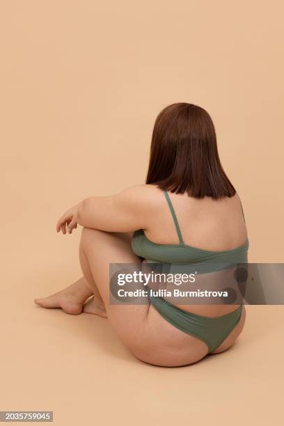 rear view of unrecognizable obese lady in bikini sitting in empty studio, hugging bent legs, exuding restlessness. - exuding stock pictures, royalty-free photos & images