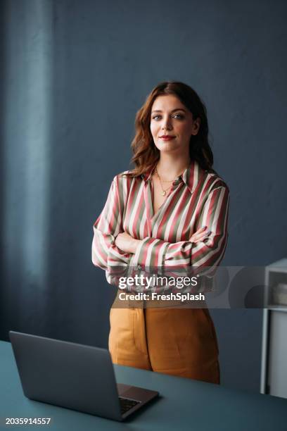 mujer de negocios segura con los brazos cruzados de pie junto a la computadora portátil - mujeres de mediana edad fotografías e imágenes de stock
