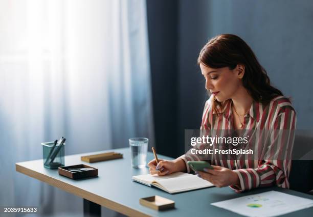 focused professional woman working at desk with notebook and smartphone - routine stock pictures, royalty-free photos & images