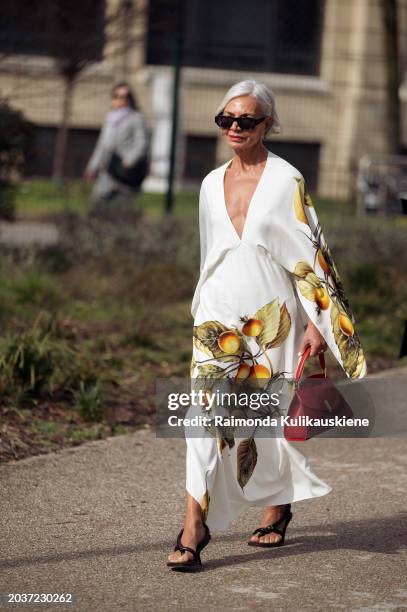 Grece Ghanem wears sunglasses, a white low-neck v-neck long flowing dress with printed orange fruits and green leaves and flared sleeves outside...