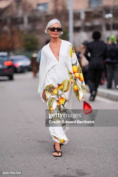 Grece Ghanem wears sunglasses, a white low-neck v-neck long flowing dress with printed orange fruits and green leaves and flared sleeves, sandals, a...