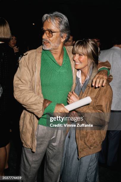 American film director and actor John Derek, wearing a beige jacket over a green sweater and grey trousers, his arm around his wife, American actress...