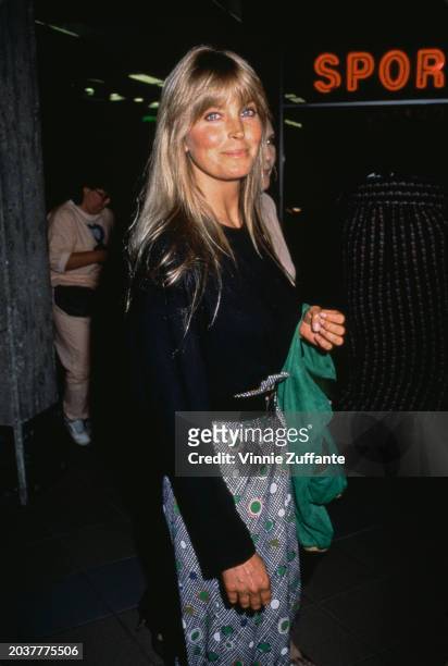American actress Bo Derek, wearing a black crew neck sweater, a green jacket over left arm, and a patterned skirt, United States, circa 1995.