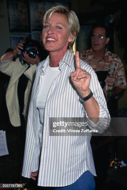 American television host and comedian Ellen DeGeneres, wearing a striped white shirt over a white t-shirt, holding up her left index finger, attends...