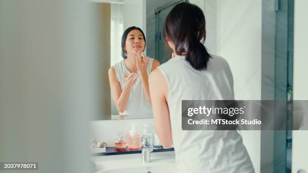 young asian woman having fun apply facial cream look in mirror in lavatory at home late morning. routine lifestyle in house. - products stock pictures, royalty-free photos & images